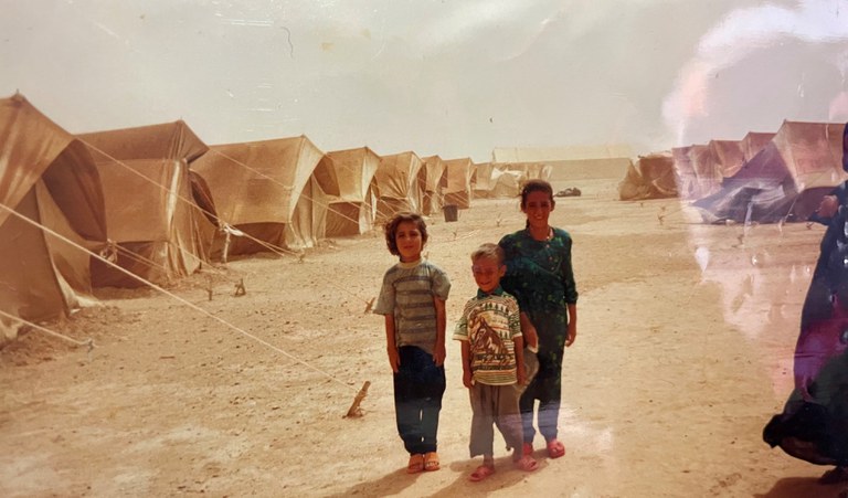 Shahan and 2 siblings standing in front of refugee camp tents.