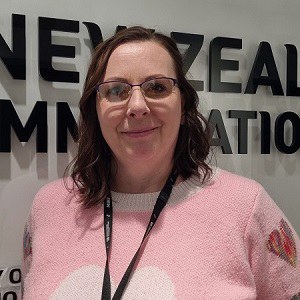 Romana stands by a white wall with text saying New Zealand Immigration